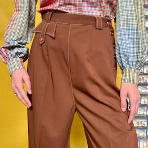 Könnte beinhalten: Braune Hose mit hohem Bund, Knopfverschluss und weißen Nahtdetails. Die Hose hat eine maßgeschneiderte Passform und ein klassisches Design. Das Model trägt ein kariertes Hemd in Grün, Rot und Blau.