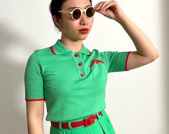 May include: A green polo shirt with red detailing, worn with matching green trousers and a red belt. The outfit is completed with cream-coloured round sunglasses.
