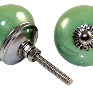 Pode incluir: Dois botões de armário de cerâmica verde com base e parafuso de metal prateado. Os botões têm um acabamento brilhante ligeiramente iridescente.