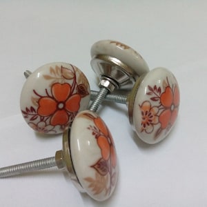 May include: Four ceramic cabinet knobs with a white background and orange floral designs. The knobs have a silver metal screw and base.