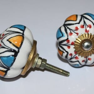 Op de afbeelding: Twee keramische deurknoppen met een witte basis en kleurrijke geometrische patronen. Eén knop heeft een gouden schroef van metaal en de andere heeft een gouden metalen basis.