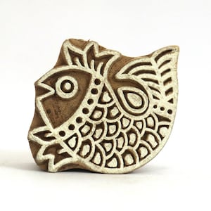 Könnte beinhalten: Ein Holzblock-Stempel mit einem detaillierten Design eines Fisches. Der Fisch hat ein großes Auge, Schuppen und Flossen. Der Stempel ist aus hellem braunem Holz geschnitzt und hat ein weißes Design.