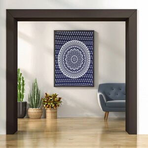 Op de afbeelding: Een witte mandala-wandtapijt met een donkerblauwe achtergrond hangt aan een witte muur in een kamer met een grijze fauteuil en planten in potten.