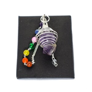 May include: Amethyst crystal pendulum in a silver wire cage with a rainbow of chakra beads on a black background.
