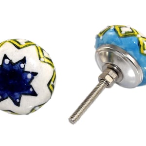 Puede incluir: Dos pomos de cerámica para armarios. Uno es blanco con un diseño de estrella azul y detalles amarillos. El otro es azul claro con patrones geométricos amarillos y blancos. Cada pomo tiene una base y un tornillo de metal plateado.