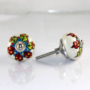 May include: Two white ceramic cabinet knobs with floral designs. One knob has a screw attached. The floral designs feature red, yellow, blue, and green colours.