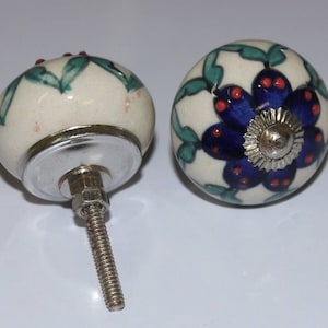 Op de afbeelding: Twee keramische deurknoppen met een witte achtergrond en bloemmotieven. De ene knop heeft een blauwe bloem met rode accenten en groene bladeren. De andere knop heeft een groen bladontwerp met rode accenten. Beide knoppen hebben een zilveren metalen basis.