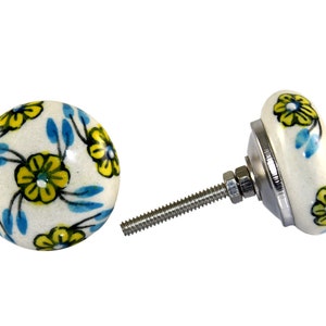 Pode incluir: Dois botões de armário de cerâmica branca com um design floral. Os botões apresentam flores amarelas com folhas azuis sobre um fundo branco. Um botão está preso a um parafuso prateado.