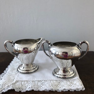 May include: A silver creamer and sugar bowl set on a white lace doily. The creamer and sugar bowl have a classic design with a rounded body and a flared base.
