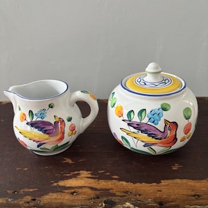 May include: A white ceramic creamer and sugar bowl set. Each piece is decorated with a colorful bird design, featuring purple, yellow, and red hues. The sugar bowl has a lid with a yellow and blue rim. The creamer has a blue rim and a handle.