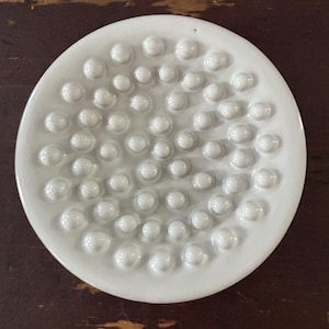 May include: A white ceramic dish with a circular shape. The dish features a pattern of raised, rounded bumps across its surface. The bumps vary in size and are evenly spaced, creating a textured appearance. The dish is set against a dark brown wooden surface.