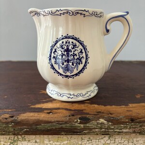 May include: A white ceramic creamer with a blue and white floral design. The creamer has a rounded body and a small spout. The handle is curved and has a blue trim.