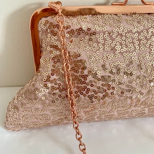 May include: A rose gold clutch purse with a chain strap and a sequined fabric. The purse has a clasp closure.