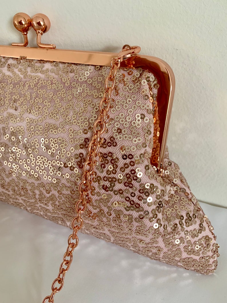 May include: A rose gold sequined clutch with a chain strap. The clutch has a clasp closure and is decorated with sequins.