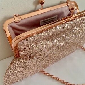 May include: A rose gold clutch purse with a chain strap. The purse is covered in sequins and has a satin lining. The purse has a clasp closure and the brand name "Glamour Purse" is printed on the inside.