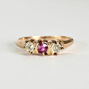 May include: A gold ring featuring a central ruby gemstone flanked by two clear diamonds. The ring has a classic design with a polished band and ornate settings for the gemstones. The gemstones are set in a prong setting.