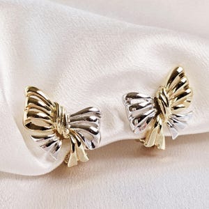 May include: A pair of gold and silver bow-shaped earrings. The earrings are made of metal and have a shiny finish.