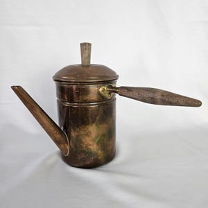 May include: A vintage copper coffee pot with a wooden handle and lid. The pot has a tarnished finish and a spout for pouring.