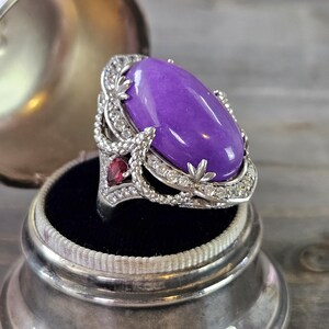 May include: A silver ring featuring a large oval purple gemstone, surrounded by smaller clear gemstones. A smaller red gemstone is set on the side of the ring.
