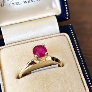 Peut inclure: Une bague en or avec une pierre précieuse ronde, facettée et d'un rouge rosé vif, montée sur quatre griffes. La bague est présentée dans un écrin bleu marine doublé de velours avec le texte J.D. Gritz, Joale St., Harrisonburg, TEL. WEN. 59301.
