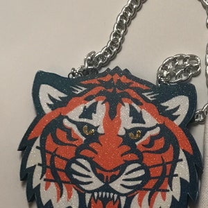 May include: A silver chain necklace with a glitter tiger head pendant. The tiger is orange, white, and blue with gold glitter eyes.