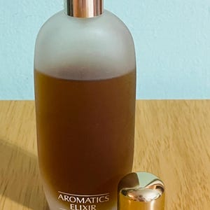 May include: A frosted glass perfume bottle with a silver cap and a separate gold-colored cap. The bottle contains a dark amber liquid and has the words "Aromatics Elixir" printed on the front.