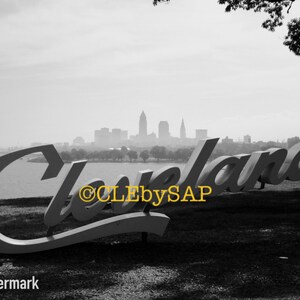 May include: A black and white photo of a large sign that reads "Cleveland" with the Cleveland skyline in the background.