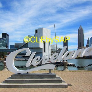 May include: A white sign with the word "Cleveland" in large, bold letters, set against a backdrop of a city skyline. The sign is positioned on a waterfront with a blue sky and white clouds.