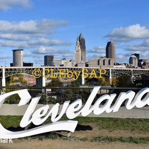 May include: A white sign with the word "Cleveland" in a large, stylized font. The sign is in front of a bridge and a cityscape with tall buildings.