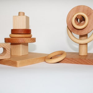 Peut inclure: Deux jouets d'empilage en bois. Le premier jouet est une tour avec une base carrée et un sommet rond. La tour a un trou au centre de la base et est en bois naturel. Le deuxième jouet est un jeu de lancer d'anneaux en bois avec une base ronde et un poteau en bois avec deux anneaux. Les anneaux sont en bois naturel et sont empilés sur le poteau. La base est en bois naturel.