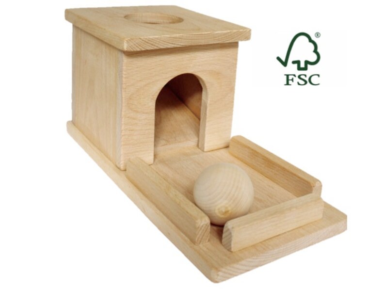 May include: A natural wood toy with a rectangular base, a small archway, and a circular opening on top. A wooden ball rests on a track in front of the structure. The FSC logo is visible.