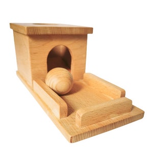 May include: A wooden toy featuring a light-colored, rectangular structure with a rounded archway and a ramp. A wooden ball rests on the ramp. The toy is designed for children's play and learning.