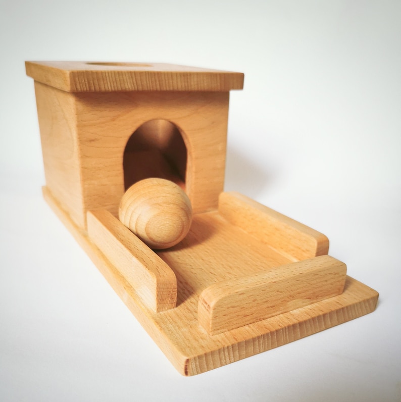 May include: A wooden toy featuring a small house with an arched doorway and a ramp. A wooden ball sits on the ramp, ready to be rolled. The toy is made of light-colored wood.