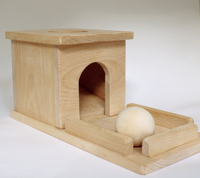 May include: A wooden toy featuring a small house with an arched doorway and a ramp. A wooden ball sits in a small enclosure in front of the house. The toy is made of light-colored wood.