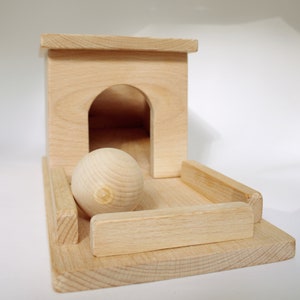May include: A wooden toy featuring a light-colored, unfinished wood design. It includes a small house-like structure with an arched doorway, a wooden ball, and a rectangular base with side rails. The toy is designed for play.