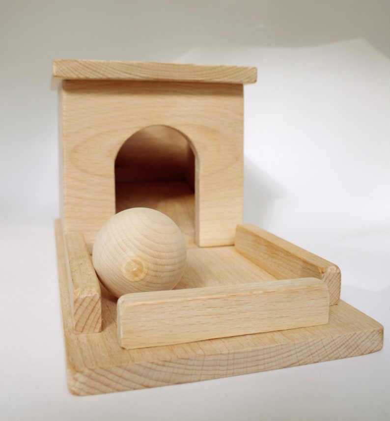 May include: A wooden toy featuring a light-colored, unfinished wood design. It includes a small house-like structure with an arched doorway, a wooden ball, and a rectangular base with side rails. The toy is designed for play.