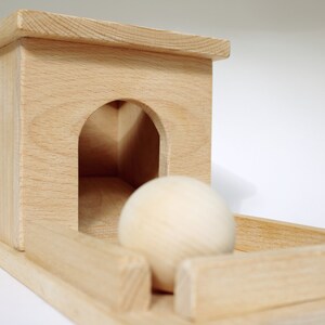 May include: A wooden toy with a light-colored finish. The toy features a small house-like structure with an arched opening and a wooden ball. The ball is positioned in front of the opening, resting on a wooden track.