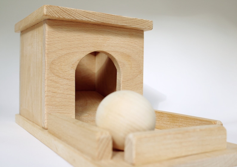 May include: A wooden toy with a light-colored finish. The toy features a small house-like structure with an arched opening and a wooden ball. The ball is positioned in front of the opening, resting on a wooden track.