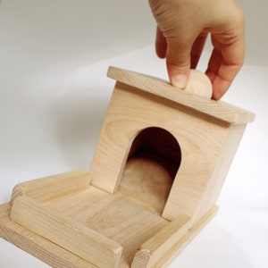 May include: A light-colored wooden birdhouse with a rounded entrance and a small wooden ball on top. The birdhouse has a rectangular base and a pitched roof. The image shows a hand touching the top of the birdhouse.