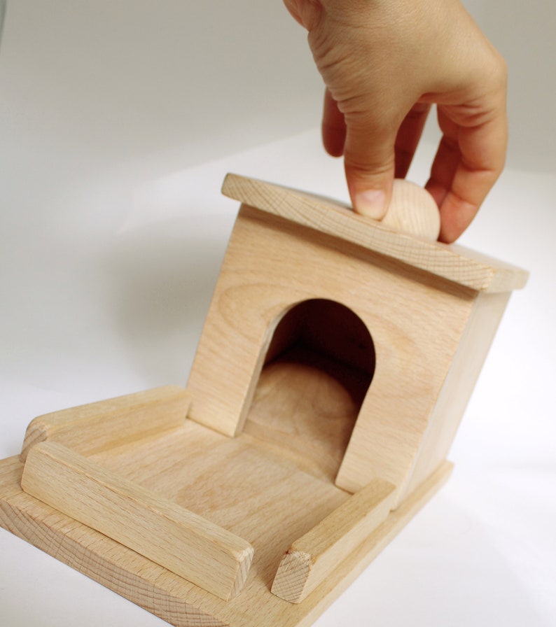 May include: A light-colored wooden birdhouse with a rounded entrance and a small wooden ball on top. The birdhouse has a rectangular base and a pitched roof. The image shows a hand touching the top of the birdhouse.
