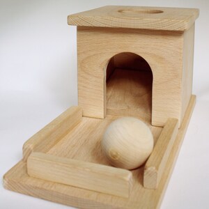 May include: A wooden toy featuring a small house with an arched doorway and a circular hole on top. A wooden ball sits on a rectangular base with raised sides. The toy is made of light-colored wood.