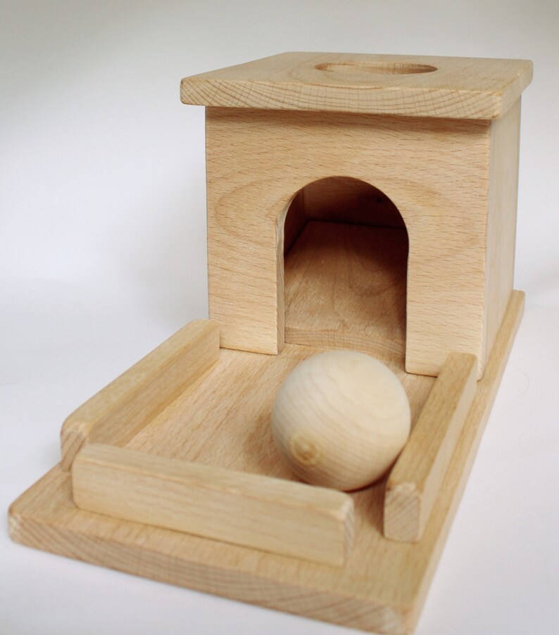 May include: A wooden toy featuring a small house with an arched doorway and a circular hole on top. A wooden ball sits on a rectangular base with raised sides. The toy is made of light-colored wood.