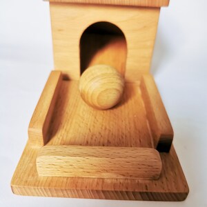 May include: A wooden toy featuring a small house with an arched doorway and a ramp. A wooden ball is positioned in the doorway, ready to roll down the ramp. The toy is made of light-colored wood.