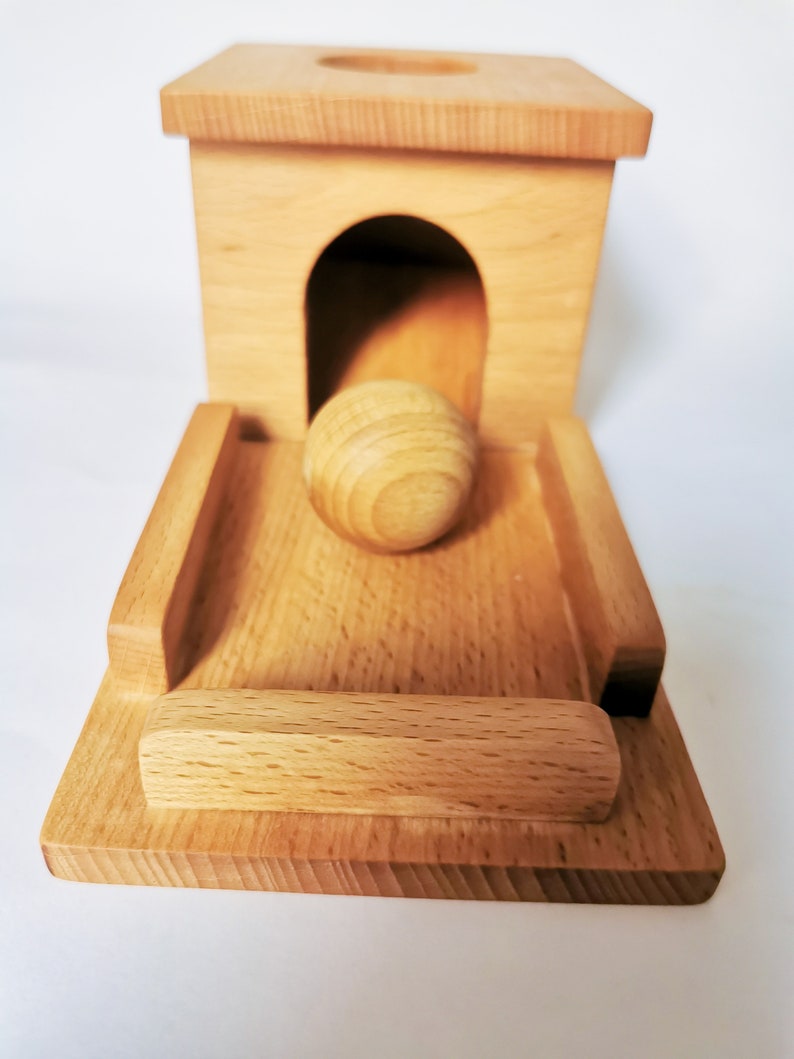 May include: A wooden toy featuring a small house with an arched doorway and a ramp. A wooden ball is positioned in the doorway, ready to roll down the ramp. The toy is made of light-colored wood.