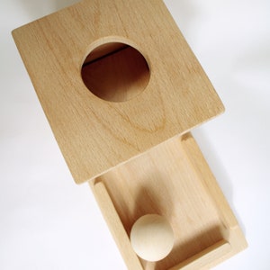 May include: A wooden toy with a square top featuring a circular hole and a rectangular drawer. A small, round wooden ball rests inside the drawer. The toy is made of light-colored wood.