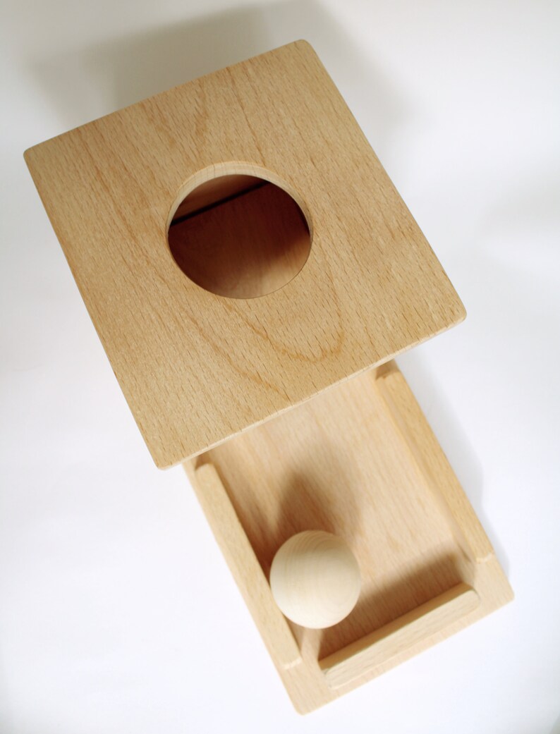 May include: A wooden toy with a square top featuring a circular hole and a rectangular drawer. A small, round wooden ball rests inside the drawer. The toy is made of light-colored wood.