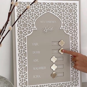 May include: A grey and white decorative wall hanging with the text "MES 5 PRIERES" and "Lylah". The design includes a decorative border and gold-coloured clover-shaped markers. The text "FAIR", "DUHR", "ASR", "MAGHREB", and "ICHA" are also visible.