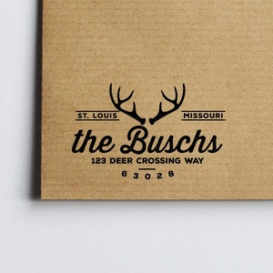 May include: A brown paper envelope with a black rubber stamp featuring a stylized deer antler logo and the text "the Buschs", "123 Deer Crossing Way", and "63028".
