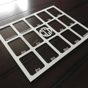 May include: White picture frame with 18 slots for photos. The slots are labeled with the grade levels from preschool to 12th grade. The center slot has a monogrammed initial.