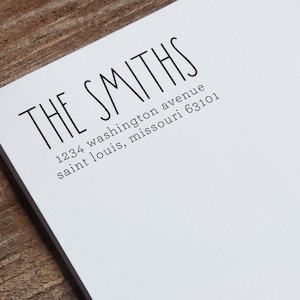 May include: White stationery with the text "THE SMITHS" in a bold, sans-serif font. Below, the address "1234 washington avenue saint louis, missouri 63101" is printed in a smaller font. The stationery rests on a wooden surface.